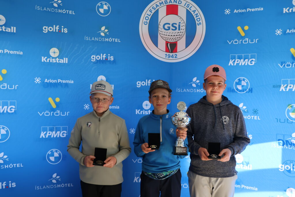 499A3990 - Golfsamband Íslands Three boys pose with trophies in front of a blue sponsor backdrop featuring logos and a large golf logo behind them.