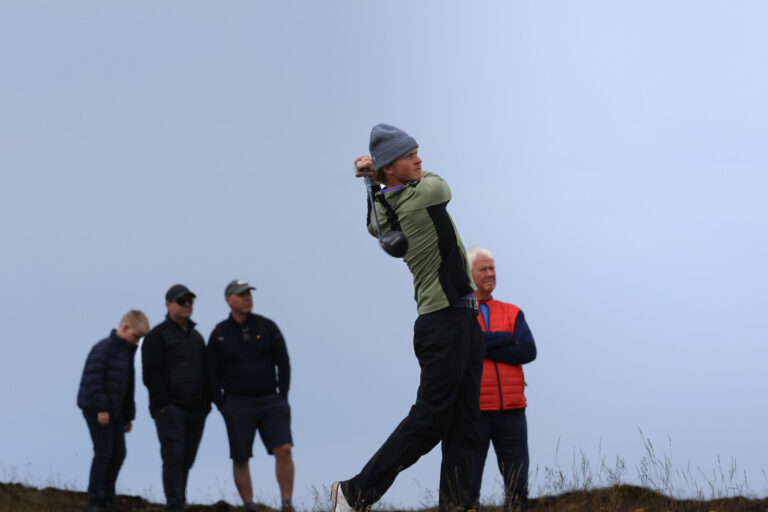 Golfer in a gray beanie and green jacket mid-swing on a grassy dune, while four spectators watch in the background.
