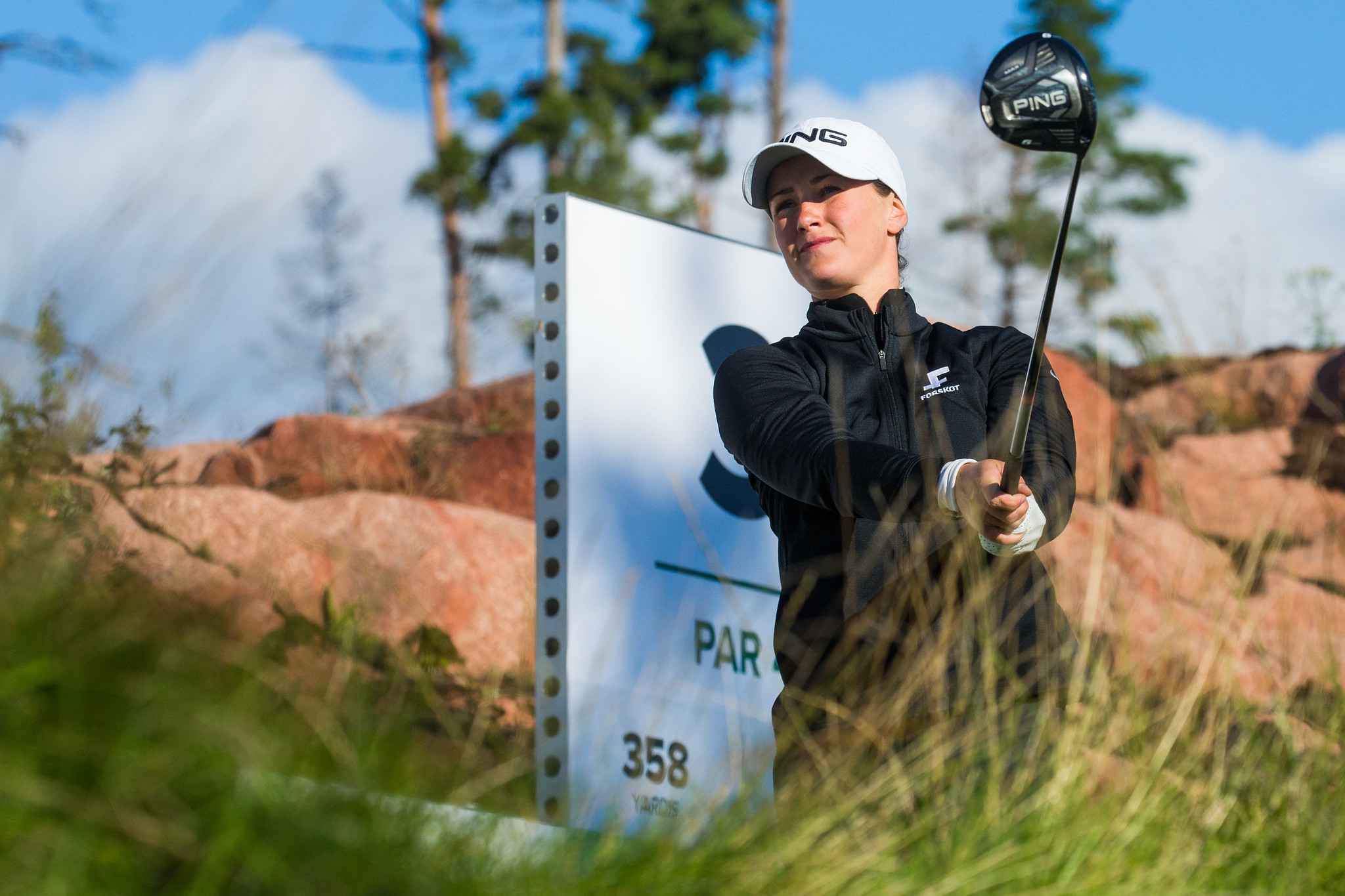 Female golfer in a black jacket and white cap prepares to swing a driver on a sunny golf course; a yardage sign shows 358 yards in the background