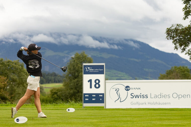 Female golfer in mid-swing on a golf course, 18th hole sign and Swiss Ladies Open banner in the background