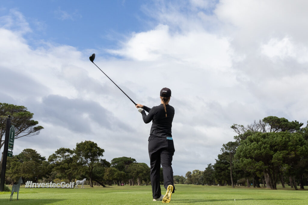 Golfer in black outfit with braided hair and yellow-soled shoes mid-swing on a lush golf course, with '#InvestecGolf' sign in the foreground.