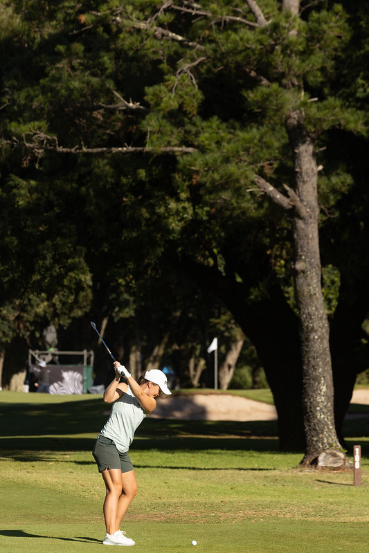 Female golfer in a white cap swings a club on a sunny golf course, ball on the grass with trees behind.