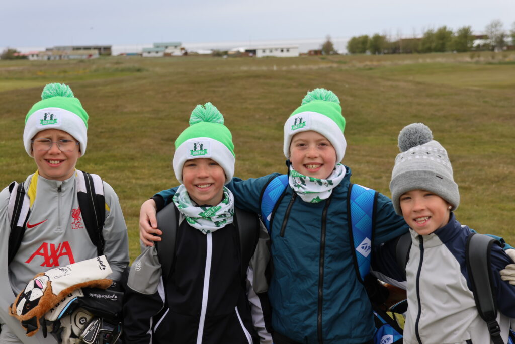 Four smiling children wearing matching green pom-pom beanies stand with their arms around each other in a grassy field.