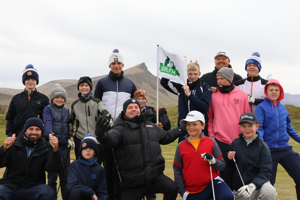 Group of young golfers posing together outdoors on a grassy hill, several wearing jackets and knit hats, a white flag reading Golf 14 visible behind them.