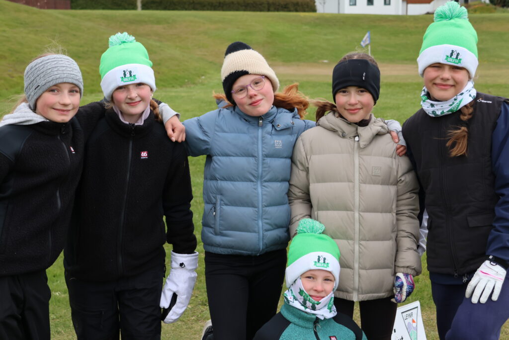 Six children stand together on a grassy field in winter jackets, hats, and gloves, smiling at the camera; a girl kneels in front wearing a white beanie with a green logo.