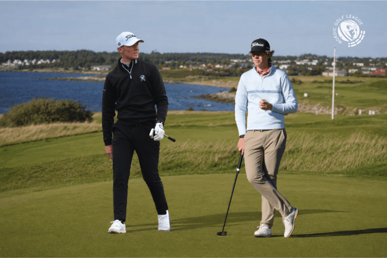 Two golfers walk on a green fairway by a coastline, club in hand; one in black, one in light blue sweater with a cap, logo visible in corner.