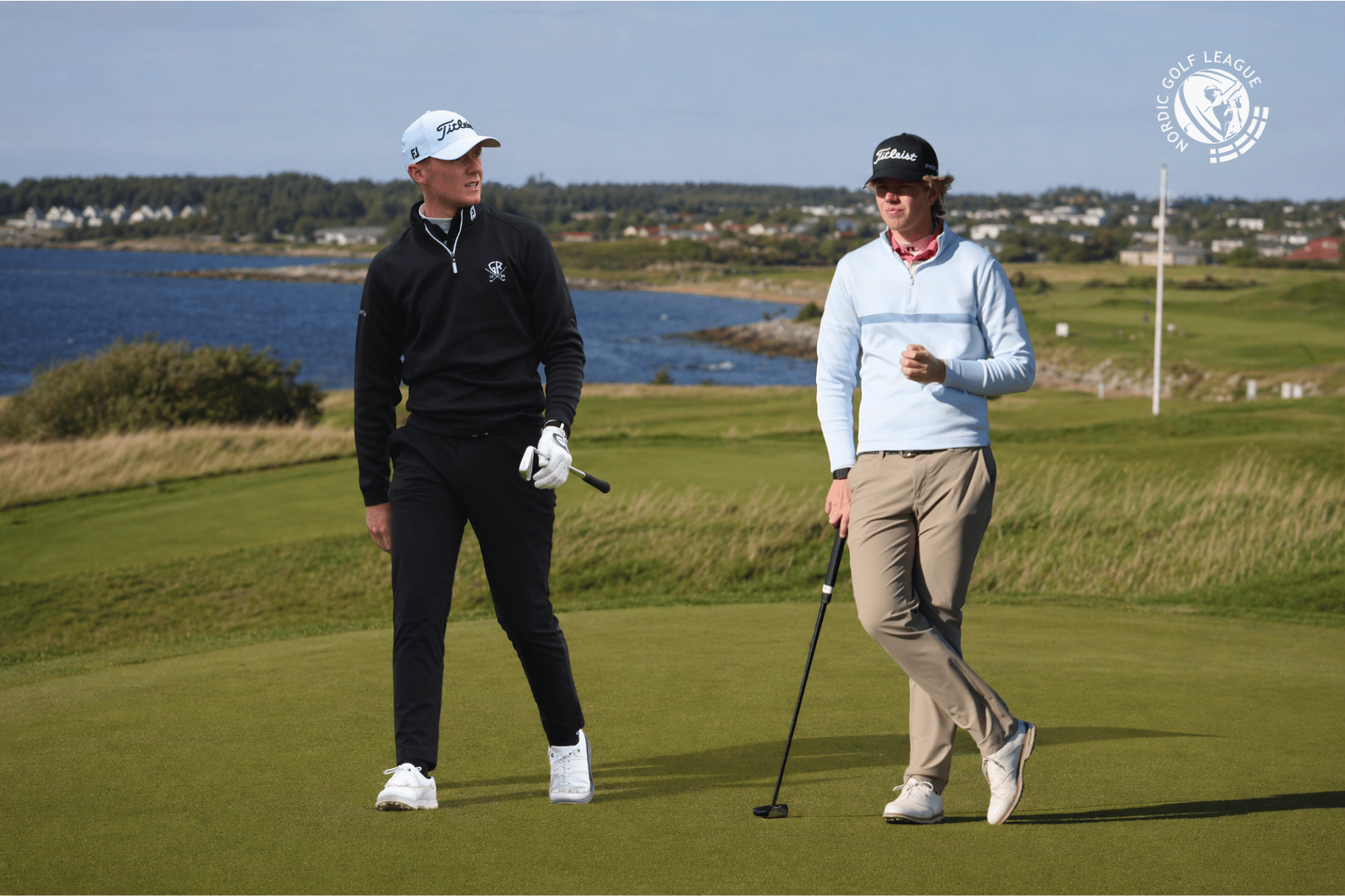 Two golfers walk on a green fairway by a coastline, club in hand; one in black, one in light blue sweater with a cap, logo visible in corner.