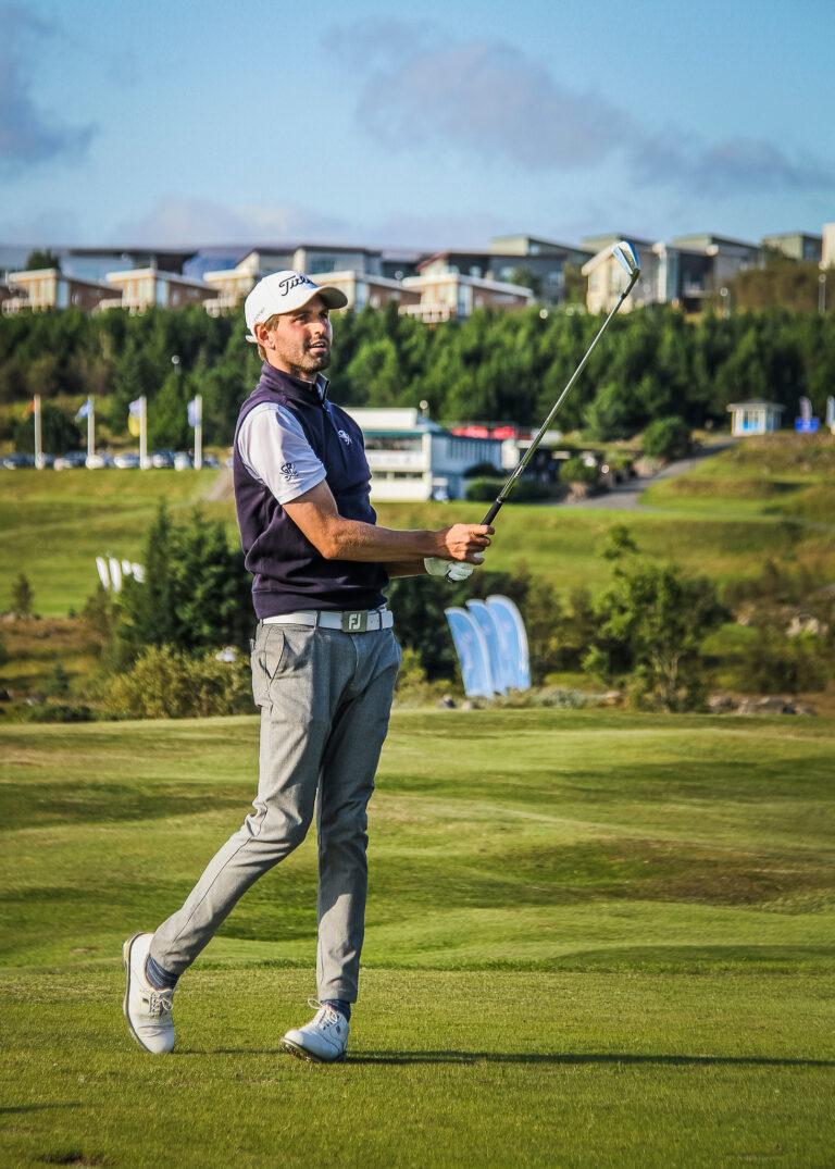 Male golfer in a navy vest and white cap swings a club on a sunny golf course with green fairways and buildings in the background.