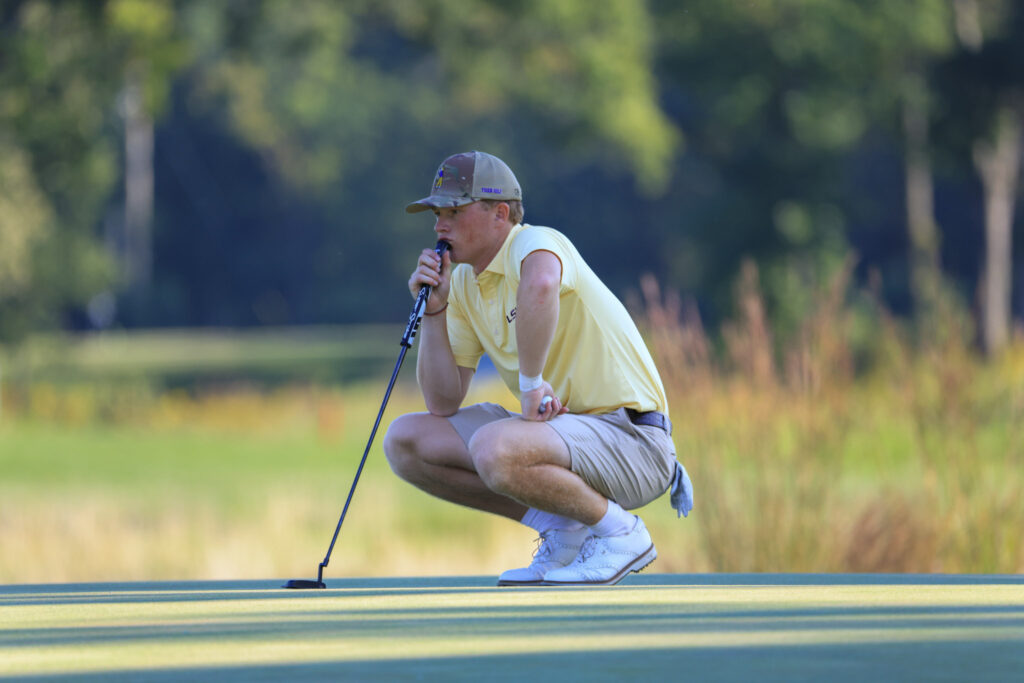 Male golfer in a pale yellow polo crouches on the green, lining up a putt with a putter.
