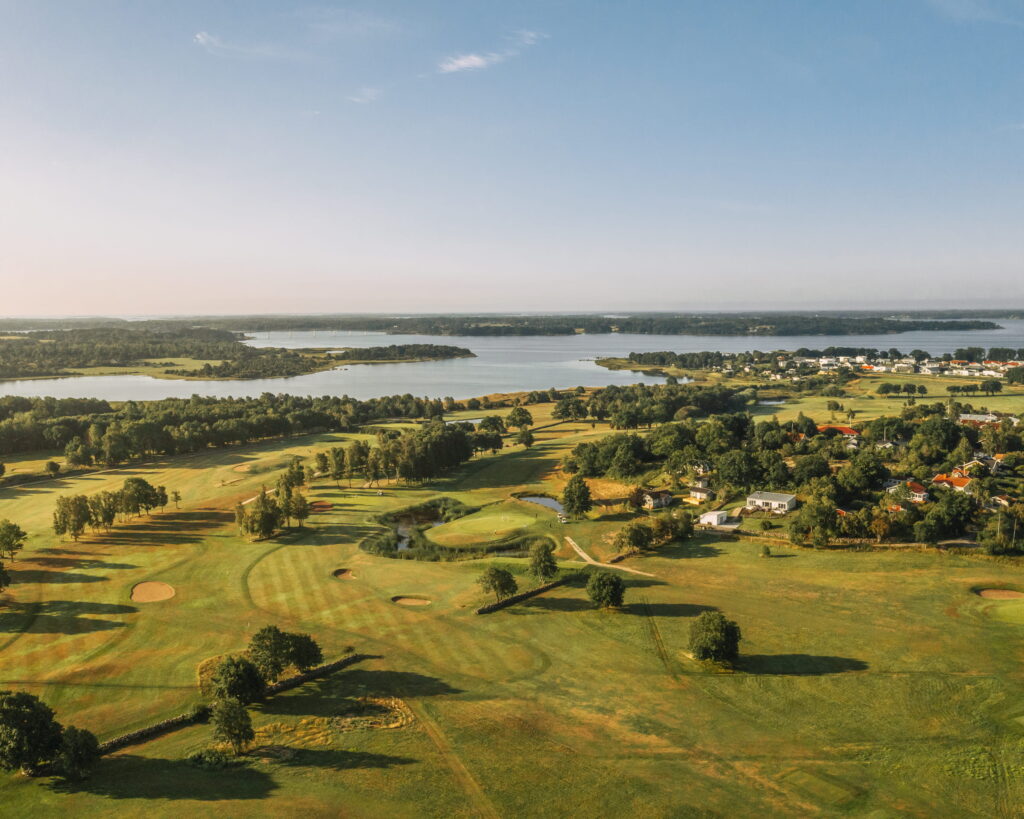 Aerial view of a golf course with green fairways, trees, a winding river, and houses along the shore under a clear blue sky at dusk light.