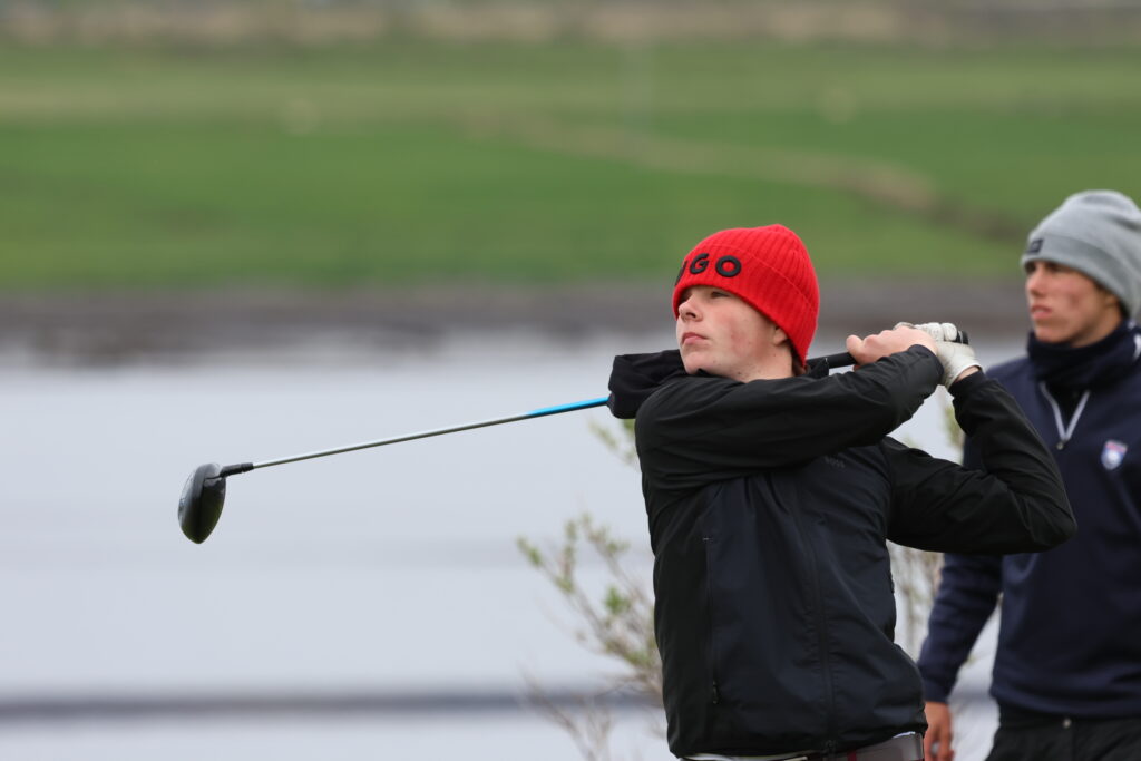 Person in a red beanie swings a golf club outdoors, with a second golfer nearby and green fields in the background.