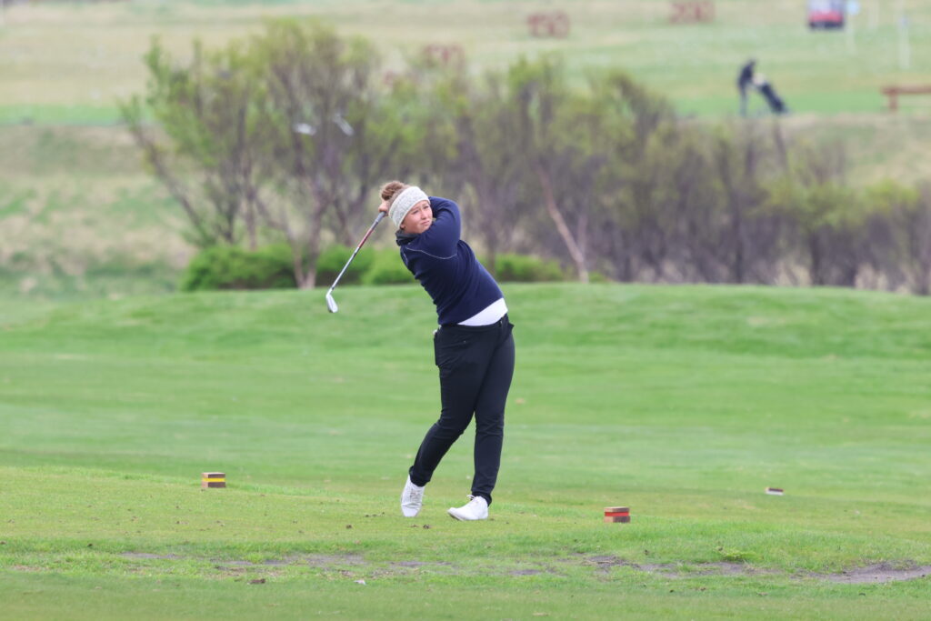 Female golfer in navy outfit swinging a club on a green golf course, with tees on the ground and trees in the background.