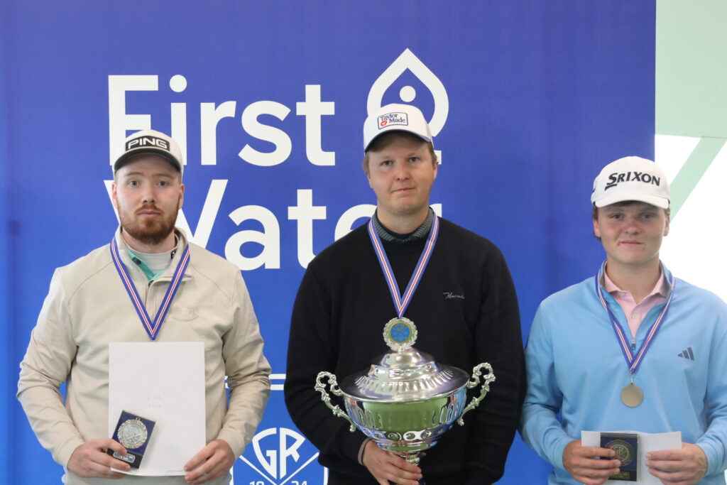 Three male athletes stand on a podium with medals around their necks; the center winner holds a large ornate trophy, while the others hold certificates and prizes against a blue backdrop.