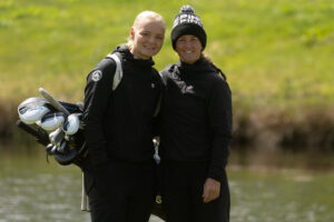 Two women in black jackets stand together outdoors, one carrying a golf bag full of clubs by a pond or lake.