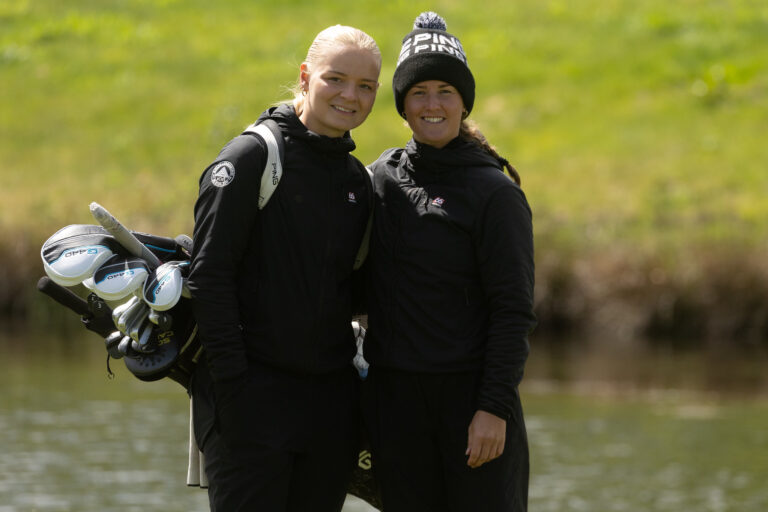 Two women in black jackets stand together outdoors, one carrying a golf bag full of clubs by a pond or lake.