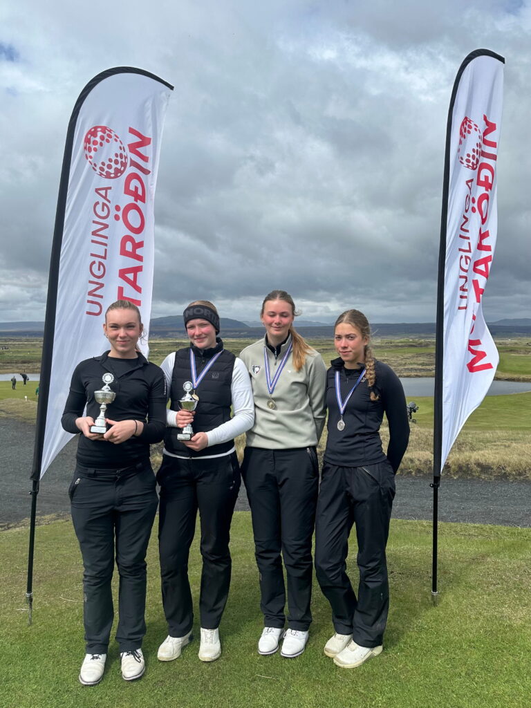 IMG_1178 - Golfsamband Íslands Four young athletes posing outdoors with medals and trophies, standing on grass with two tall white banners in the background.