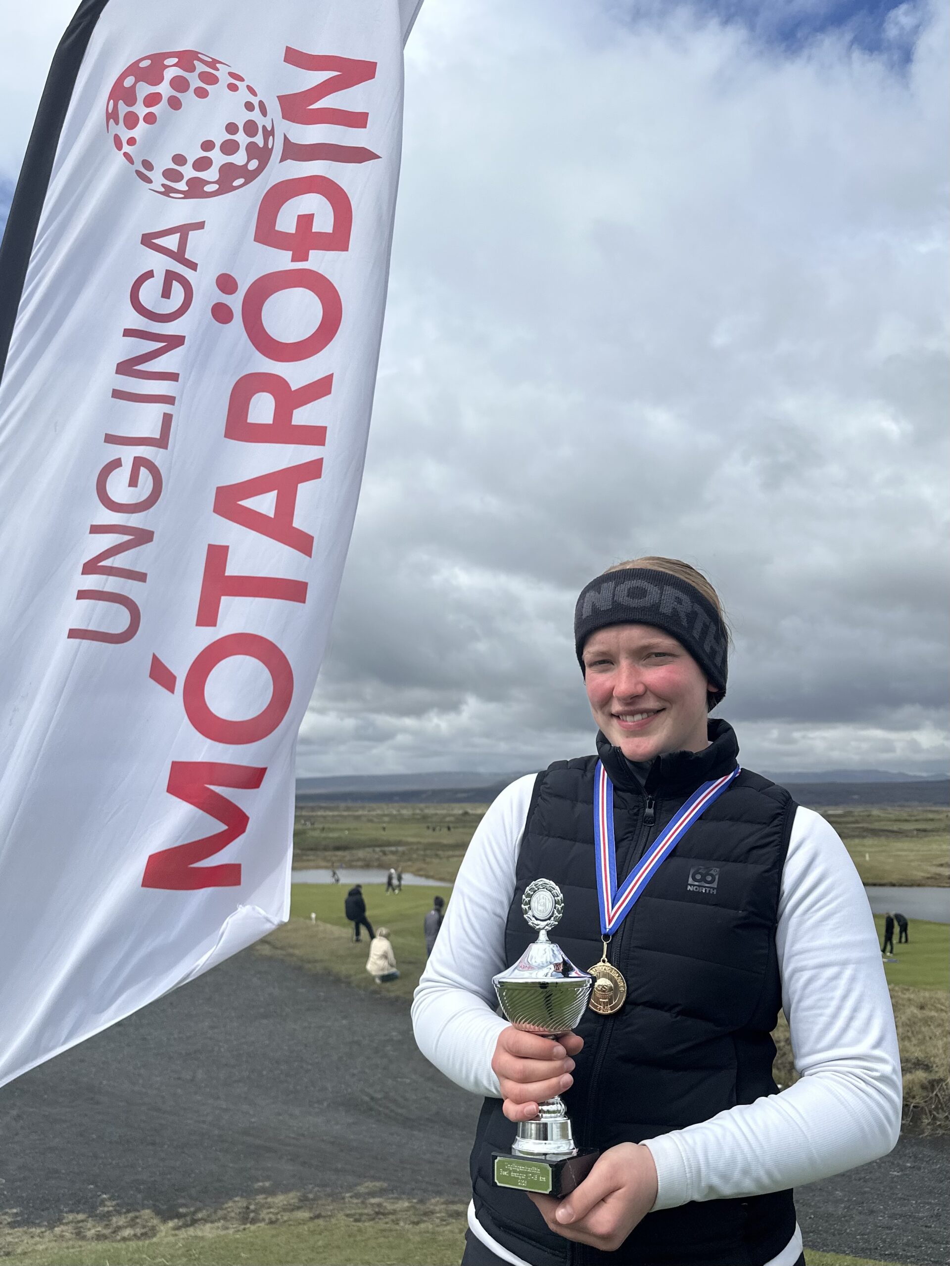 Young atleta holding a trophy and medals at an outdoor award event, standing beside a white flag with red text and logo against a cloudy sky.