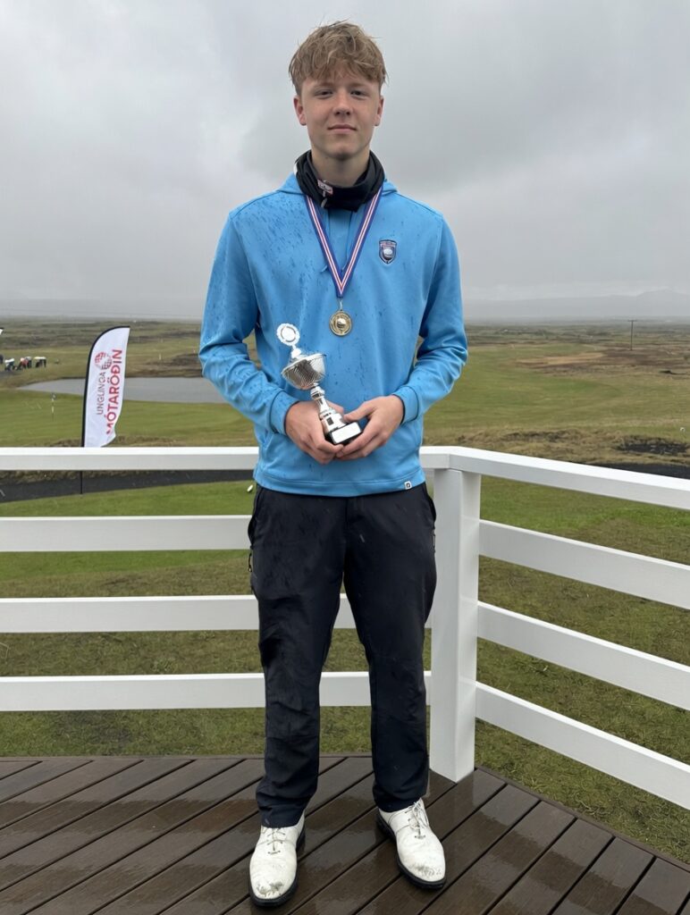 IMG_1220 - Golfsamband Íslands Young athlete standing on a wooden deck in the rain, wearing a blue jacket and black pants, holding a trophy and wearing a gold medal around the neck.