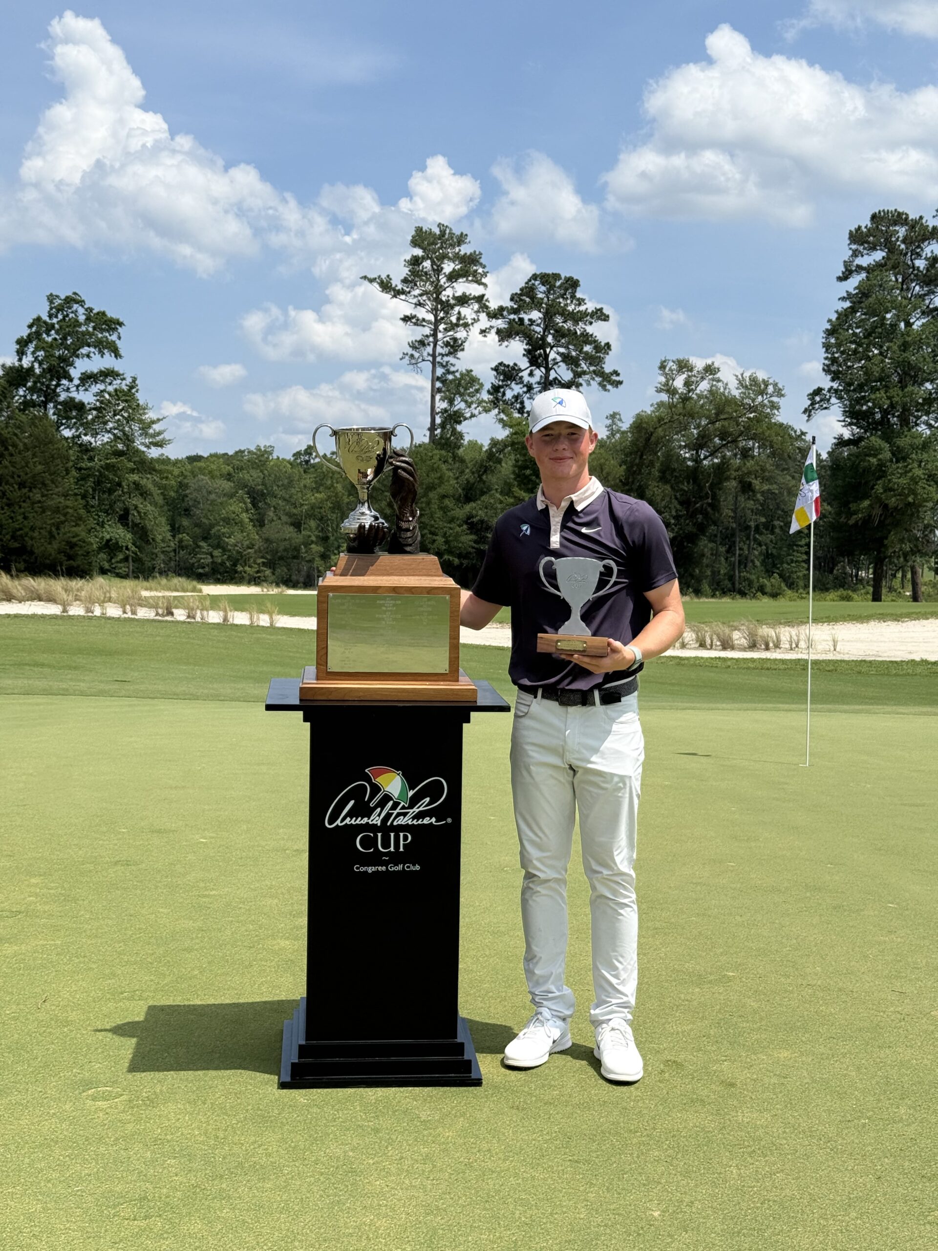 Young golfer in a dark polo and white cap poses with a large trophy on a wooden podium at a golf course, with a flag in the background and sunny skies.