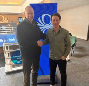 Two men shake hands indoors, standing in front of a large blue screen; one wearing a dark jacket and brown shoes, the other in a green button‑down shirt and white sneakers, both smiling.