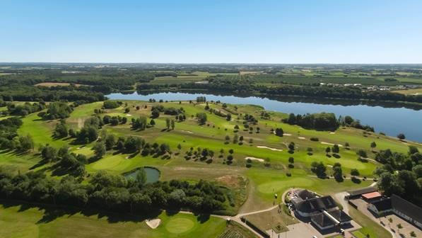 Aerial view of a lush golf course with greens, fairways, water hazards, and a clubhouse in the corner under a blue sky.