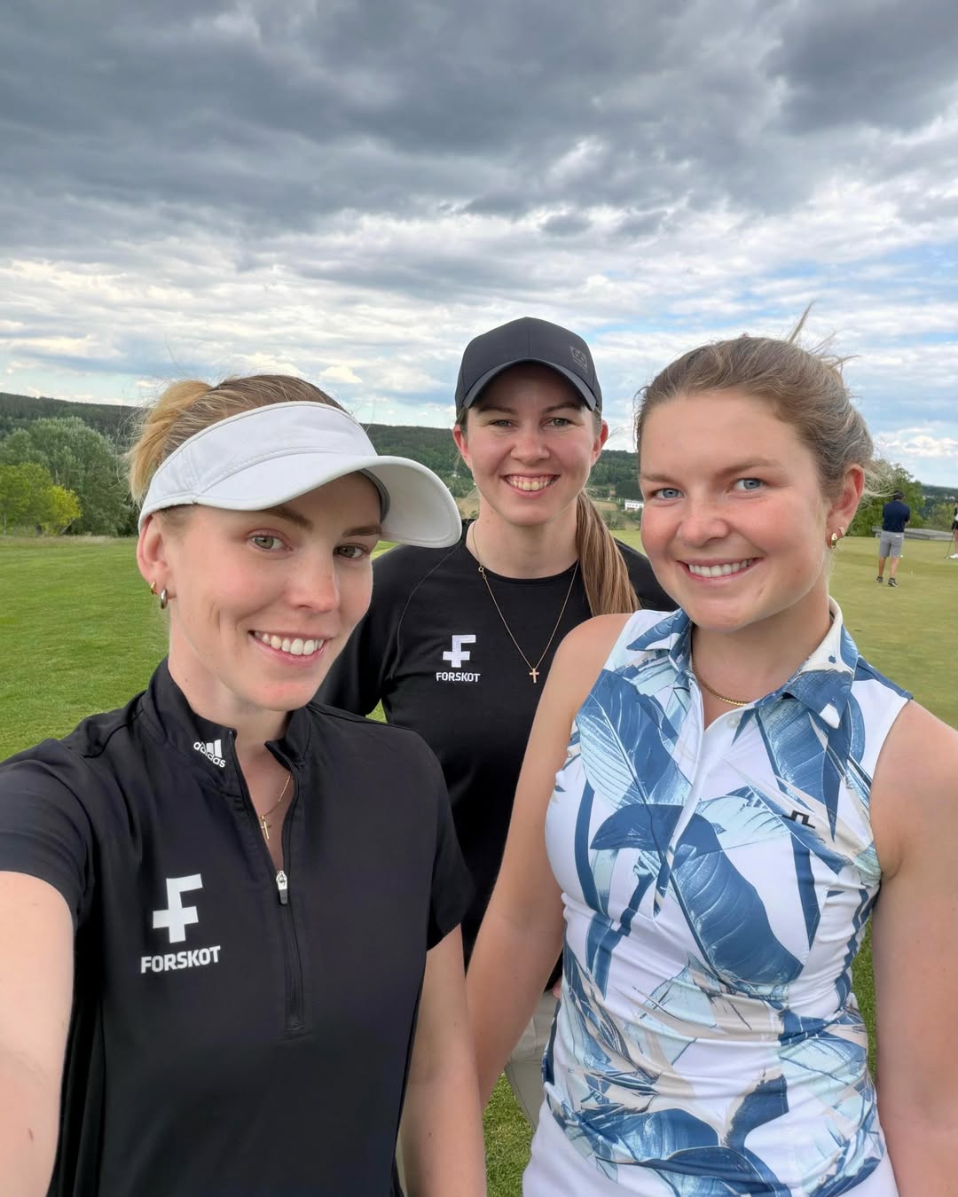 Three women taking a daytime selfie on a grassy field; two wear FORSKOT branded sportswear, one in a white visor and another in a black cap, with a cloudy sky behind them.