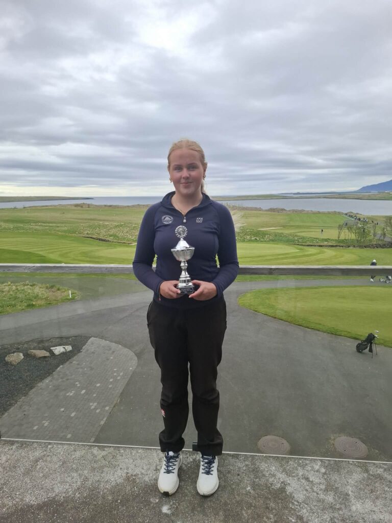 Young woman on a golf course holding a silver trophy, with a cloudy sky and green fairways in the background.