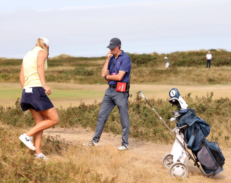 A man in a blue polo and cap stands with a red pouch at his hip, talking on a golf course as a woman in a yellow top and navy skirt jogs by; a golf bag on a wheeled cart sits in the foreground.