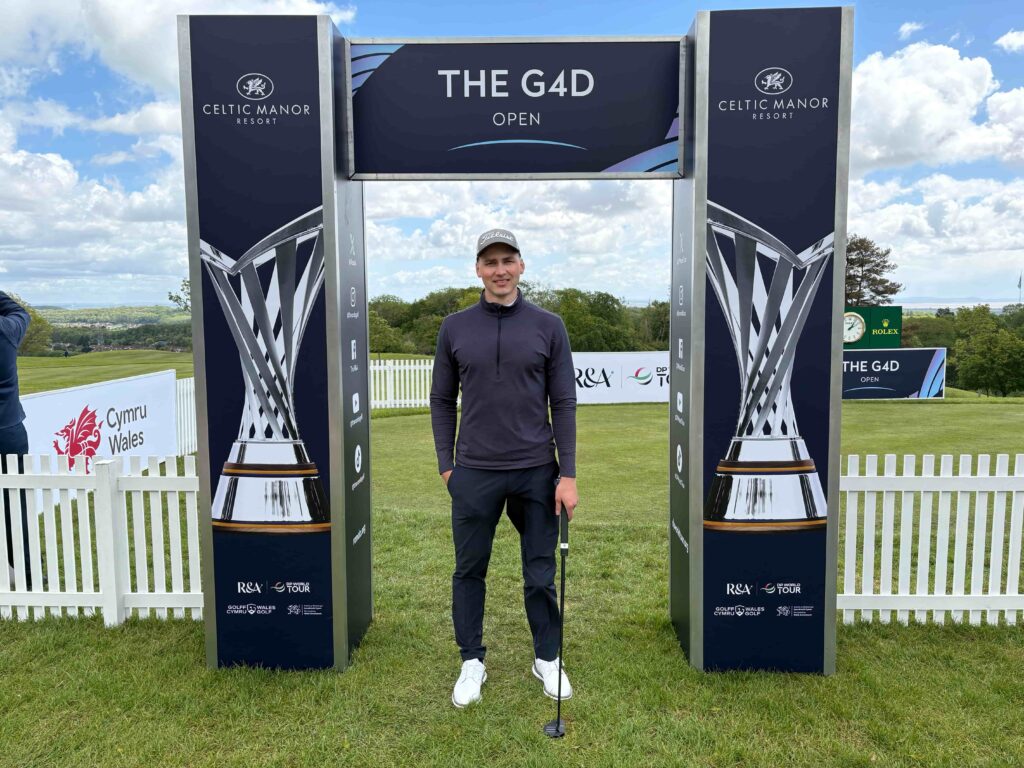 Man standing with a golf club under a dark blue ‘THE G4D OPEN’ arch at a Celtic Manor golf event