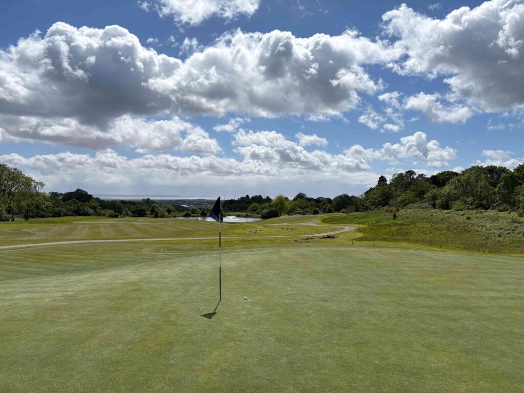 Golf green with the hole flag in the foreground, rolling fairway and trees to the right under a blue, cloudy sky.