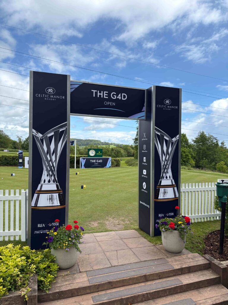 Entrance arch for The G4D Open at Celtic Manor Resort with dark blue pillars and a white picket fence, town-green in background.