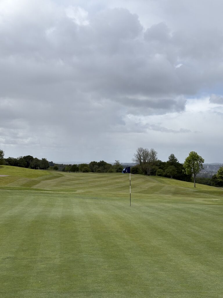 Golf course green with a flagstick showing hole number 2, surrounded by rolling fairways and distant trees under a cloudy sky.