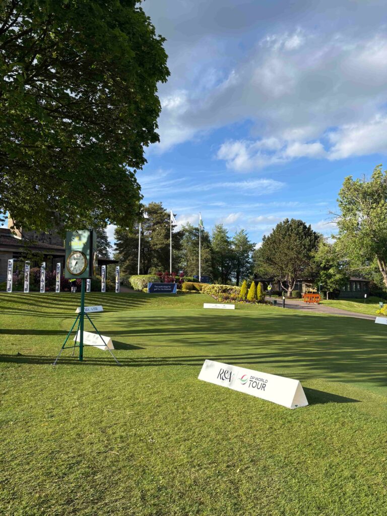 Outdoor golf-tournament display on a manicured lawn with a large Rolex clock sign and flags under a blue sky with clouds.