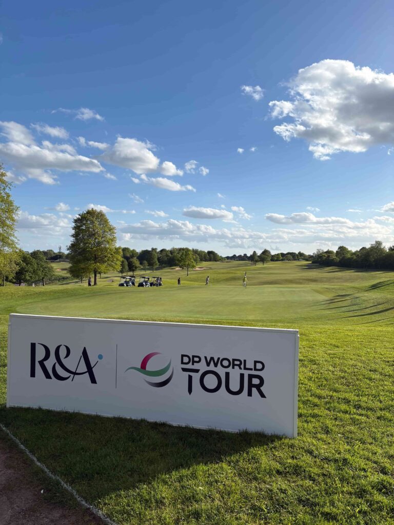 Golf course with a large DP World Tour sign in the foreground and green fairways under a blue sky with scattered clouds.