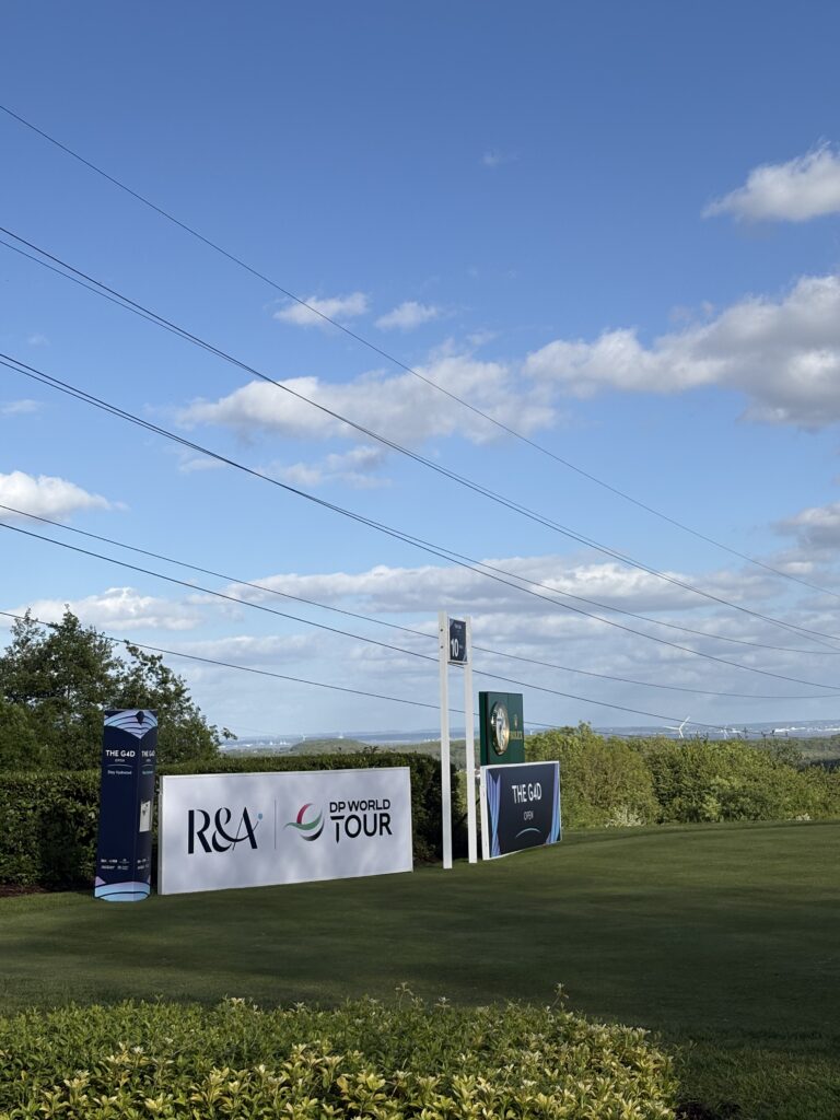 Event advertising banners for a golf tournament on a grassy field with a clear blue sky and overhead power lines.