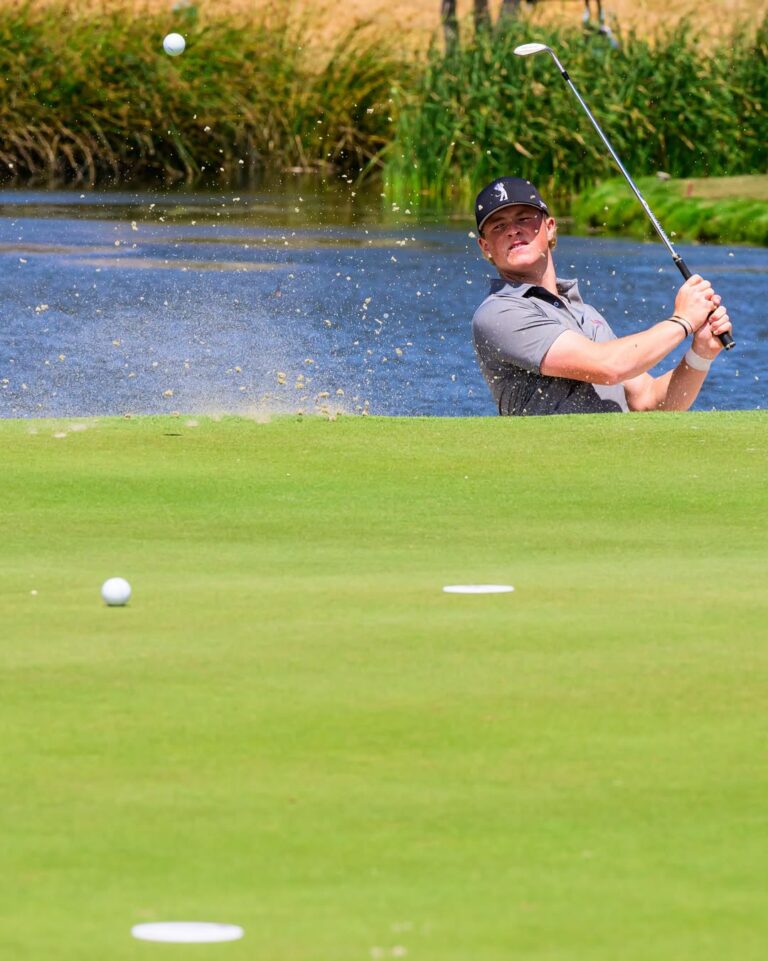 Male golfer in a gray polo and cap hits a shot from a sand bunker with water behind, ball rising through the air.
