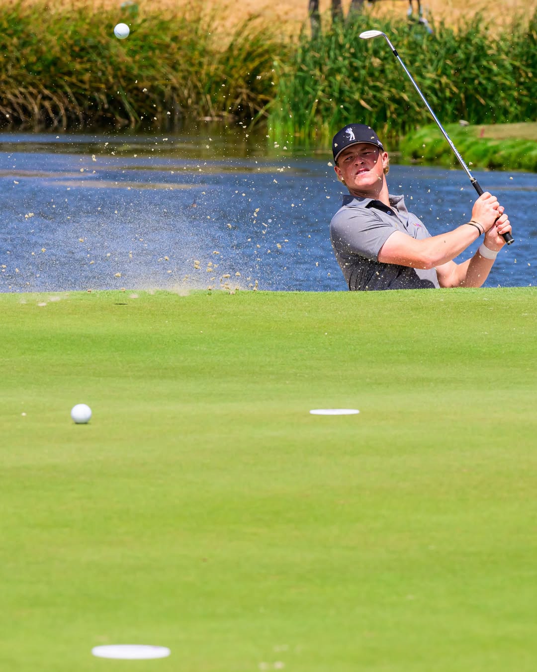 Male golfer in a gray polo and cap hits a shot from a sand bunker with water behind, ball rising through the air.