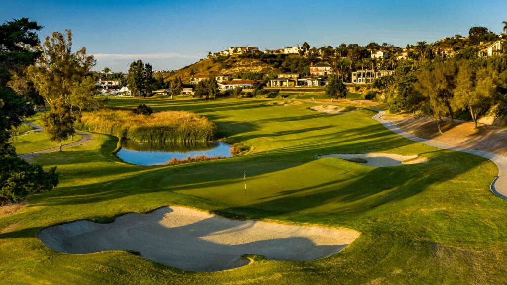Sprawling golf course with greens, sand bunkers, a water hazard, and hillside houses in the distance under a blue sky.