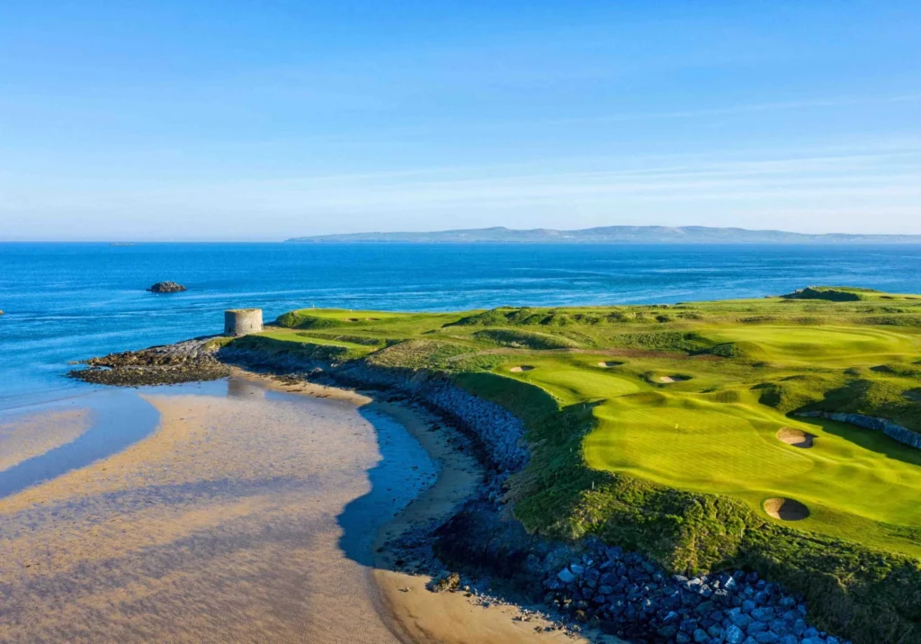 Coastal landscape with a blue sea, clear sky, rocky shoreline, and a small stone turret on the grassy coast.
