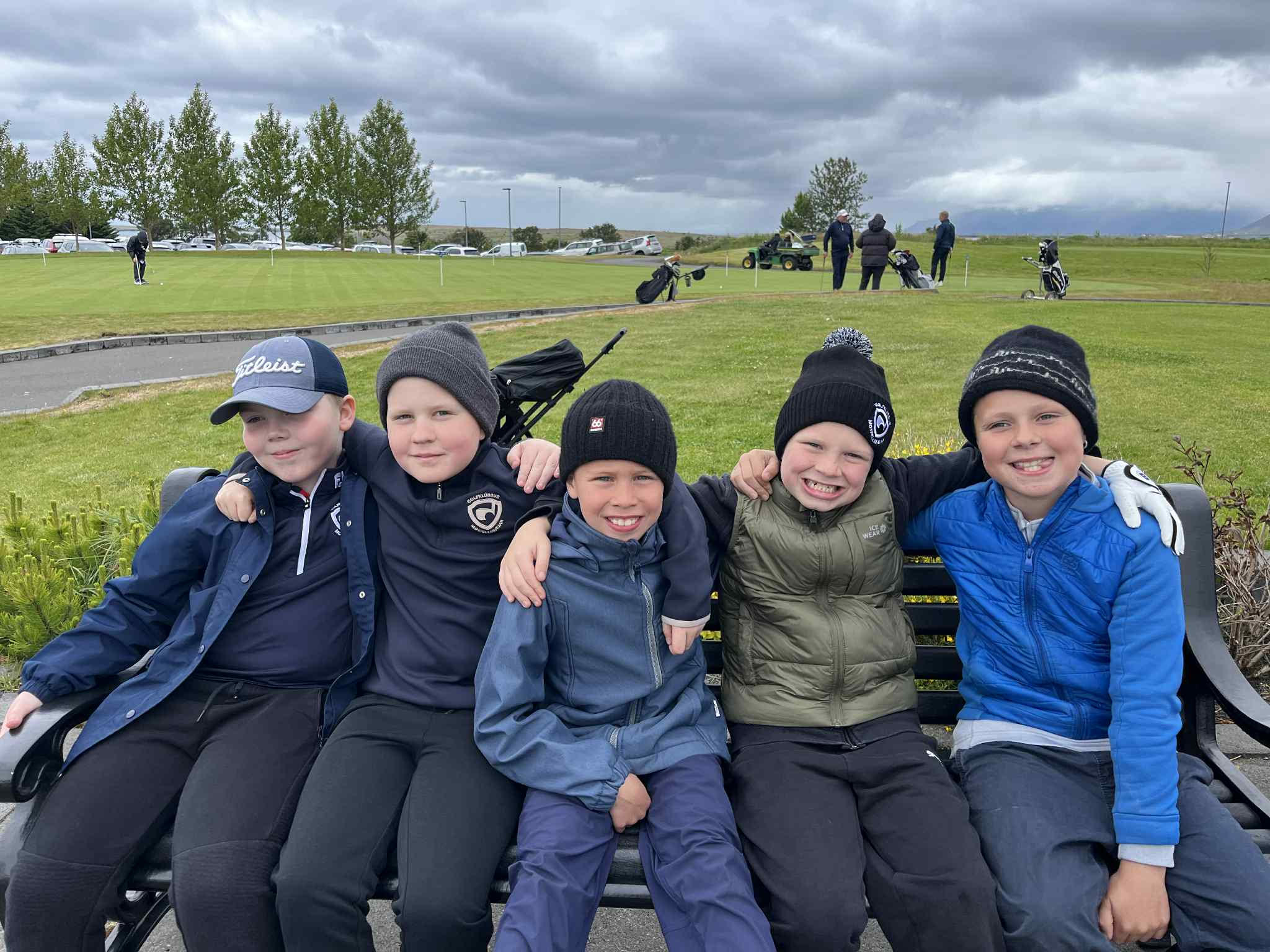 Five children in jackets sit close together on a bench at a golf course, with players and carts in the background under a cloudy sky.