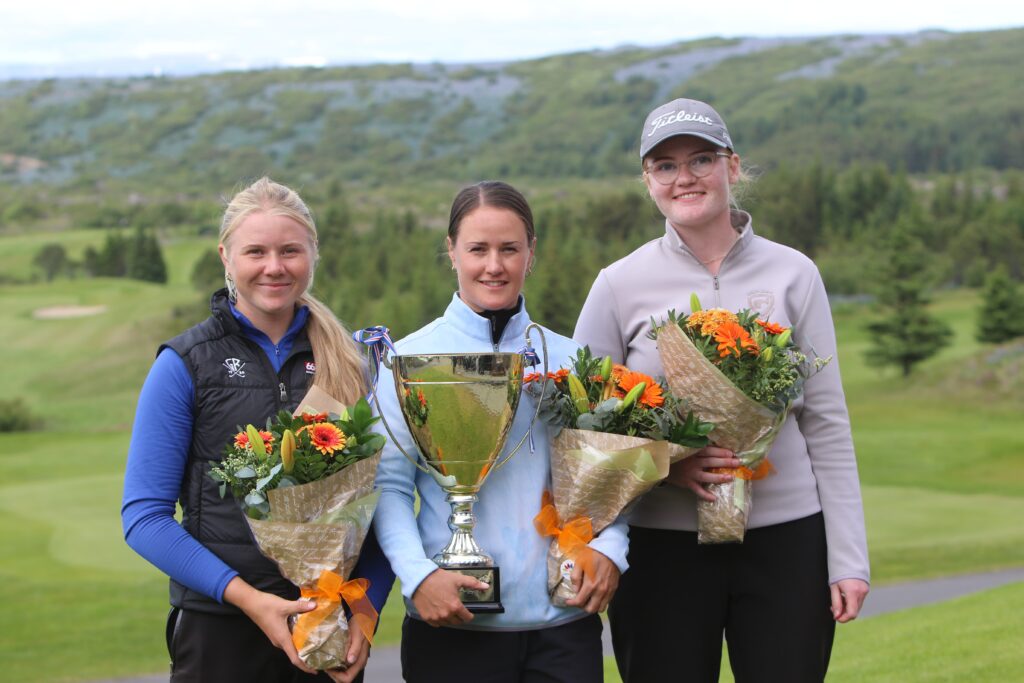 088A0314 1 - Golfsamband Íslands Three women stand outdoors, holding a large trophy and bouquets, smiling at the camera. a scenic green landscape behind them.