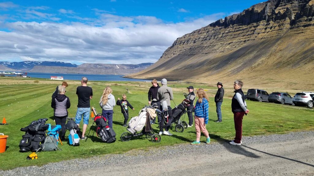 1000023895 1 - Golfsamband Íslands Group of people with golf bags and push carts on a grassy course, mountains in the background and parked cars nearby by the coast.