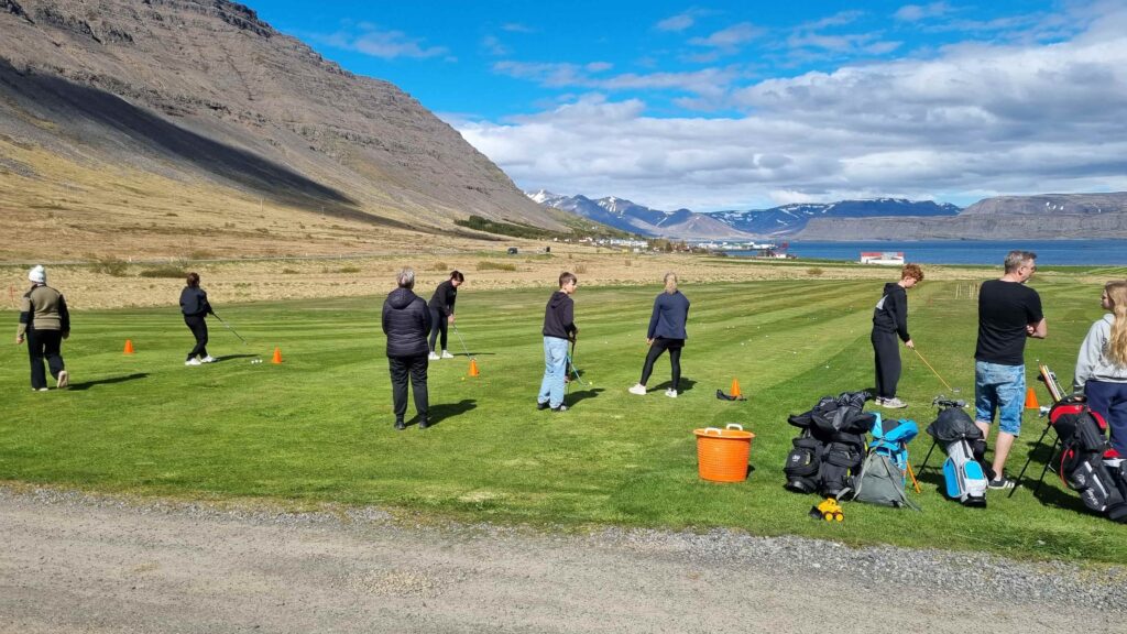 1000023896 1 - Golfsamband Íslands Group of people practicing golf on a grassy field with bags and clubs, orange cones on the ground, mountains and a lake in the background.
