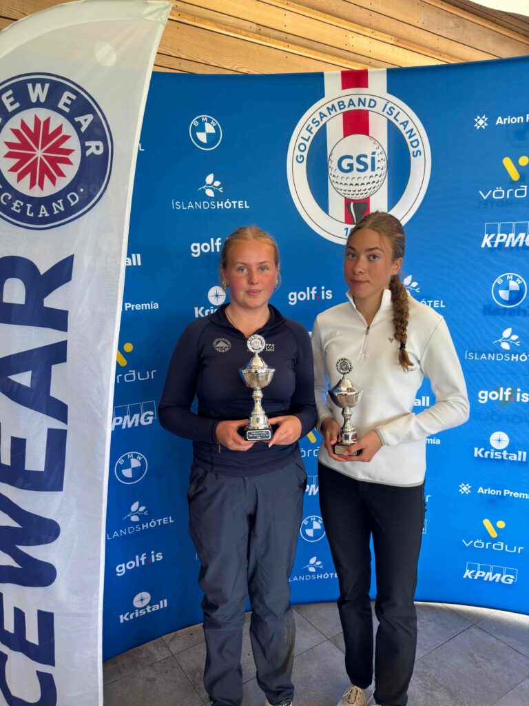 Two young athletes stand in front of a blue event backdrop, each holding a trophy after a competition.