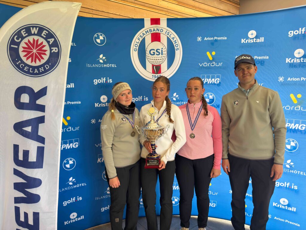 Three young athletes pose with medals and a trophy at a sports award ceremony, in front of a blue sponsor backdrop and a man in a cap nearby.