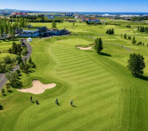 Aerial view of a sunny golf course with green fairways, sand bunkers, and a clubhouse in the distance near the coast.