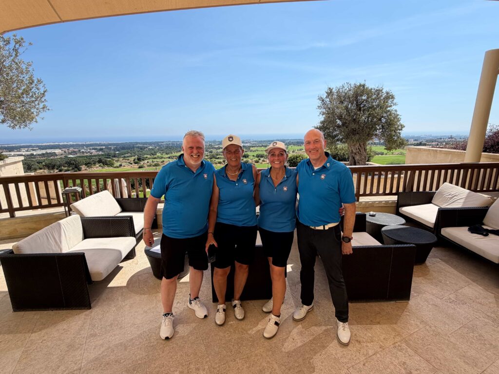 Four adults in matching blue polo shirts smile and pose on a terrace with a coastal landscape and blue sky behind them.