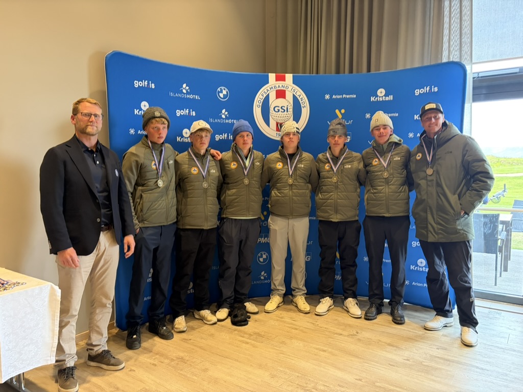 Group of eight young athletes wearing medals and gray jackets pose with two adults in front of a blue sponsor backdrop at an awards event.
