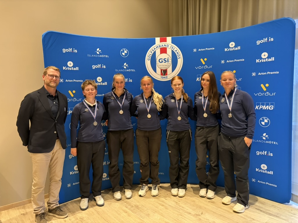 Group of seven people—six young female athletes and one male coach—standing with medals in front of a blue sponsor backdrop.