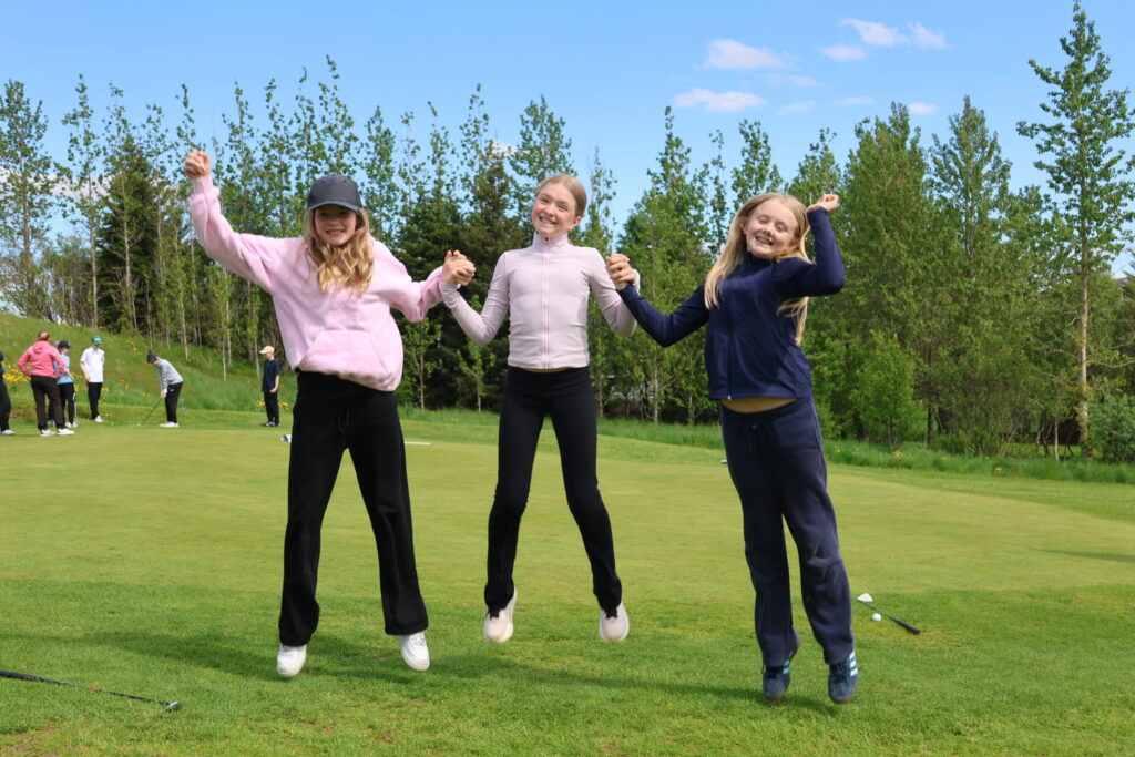 499A0342 - Golfsamband Íslands Three smiling girls jump holding hands on a sunny golf course, with other players in the background.